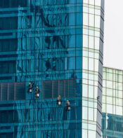 Trabajadores de limpieza en el exterior de un rascacielos de vidrio azul, utilizando cuerdas y arneses para acceder a las ventanas. Reflejos de la estructura en la superficie del edificio.